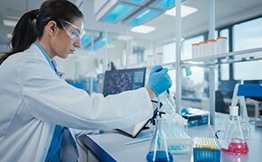 Female lab worker wearing safety goggles and nitrile gloves prepares solution of reagents in beakers with blue and pink liquid.