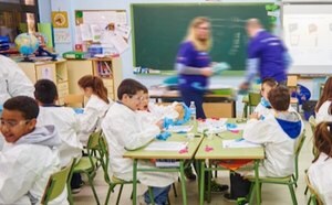 Un grupo de estudiantes sentados en sus mesas del colegio ataviados con batas de laboratorio y guantes. Dos voluntarios del personal se pasean entre las mesas para ayudar a los estudiantes.