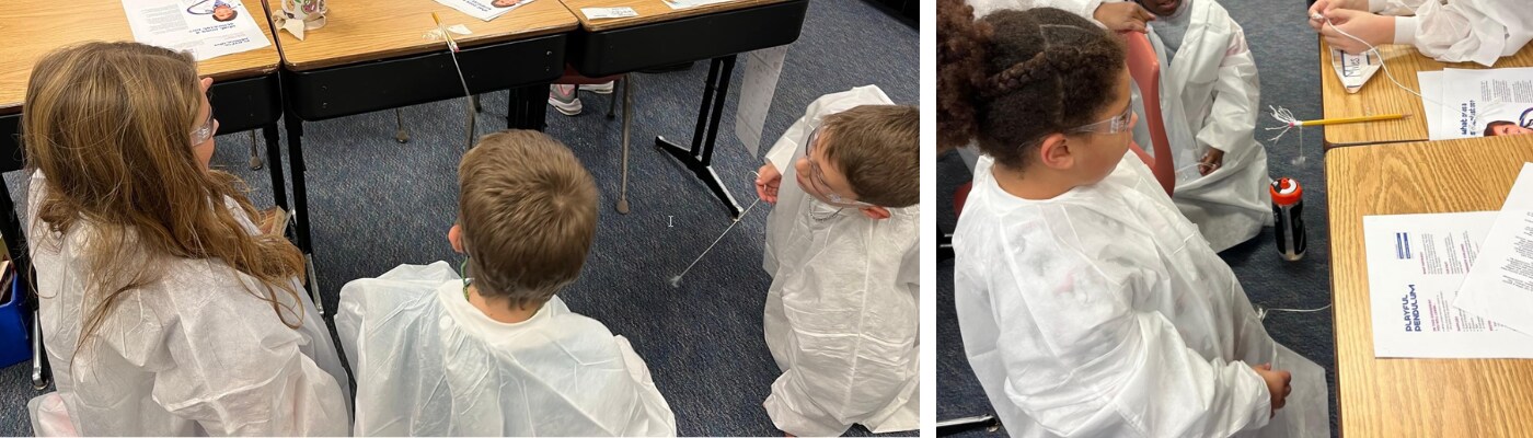 Students wear safety glasses and lab coats while watching a pendulum swing from a desk.