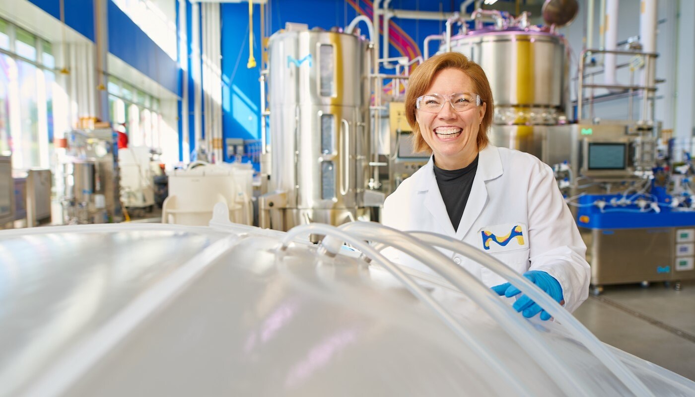 Shannon lleva una bata de laboratorio, guantes azules y gafas de seguridad y est&aacute; de pie en un M Lab: un entorno de fabricaci&oacute;n biol&oacute;gica no GMP. Ella est&aacute; sonriendo y sosteniendo una bolsa de basura, que est&aacute; situada en la esquina inferior izquierda.