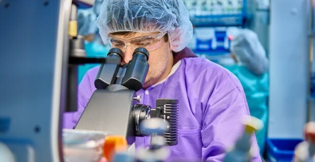 Los cient&iacute;ficos llevan una bata de laboratorio morada, gafas protectoras y una redecilla del pelo mientras miran con detenimiento en un microscopio.